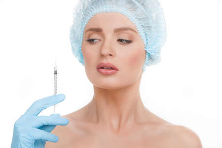 Fear of injections. Portrait of terrified young woman in medical headwear looking at the hand holding syringe near her face isolated on whiteの写真素材