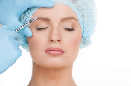 Injection. Portrait of beautiful young woman in medical headwear keeping eyes closed while doctors hands in gloves making a botox injection in her face isolated on whiteの写真素材