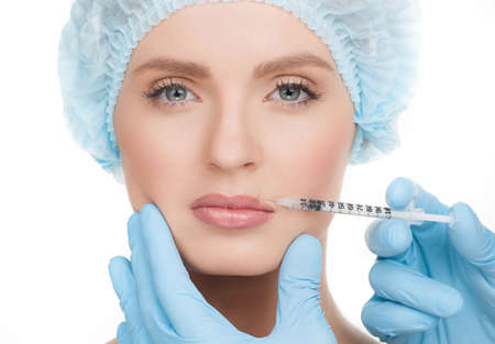 Botox injection. Portrait of beautiful young woman in medical headwear looking at camera while doctors hands in gloves making a botox injection in her face isolated on whiteの写真素材