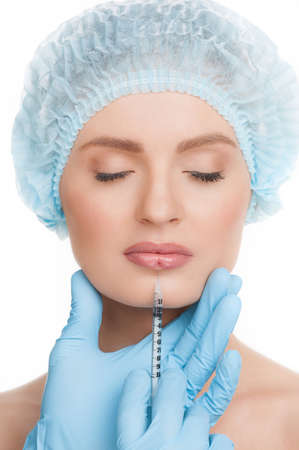Botox injection. Portrait of beautiful young woman in medical headwear keeping eyes closed while doctors hands in gloves making a botox injection in her lipsの写真素材