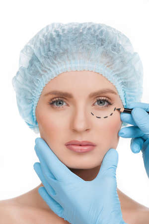Making marks on face. Portrait of beautiful young woman in medical headwear looking at camera while doctors hands in gloves making marks on her face isolated on whiteの写真素材
