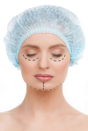 Before plastic surgery. Beautiful young woman in medical headwear and markings on face keeping her eyes closed while isolated on whiteの写真素材
