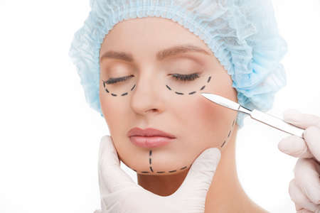 Scalpel near face. Portrait of beautiful young woman in medical headwear keeping eyes closed while doctors hands in gloves holding scalpel near her faceの写真素材
