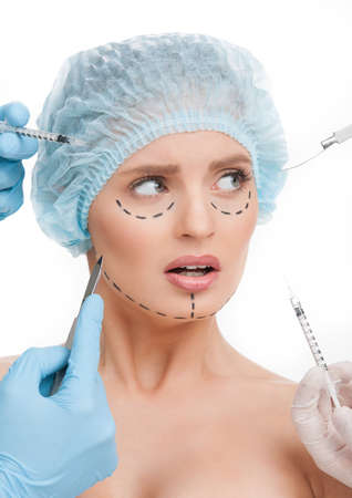 Plastic surgery. Terrified young woman looking away while many hands holding syringes and scalpels near her faceの写真素材