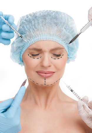 Plastic surgery. Terrified young woman keeping eyes closed while many hands holding syringes and scalpels near her faceの写真素材