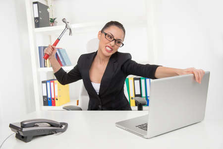 Stressed businesswoman. Angry young businesswoman trying to break her laptop with hummerの写真素材