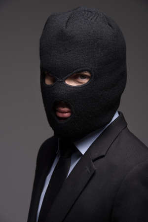 White collar crime. Portrait of businessman in black balaclava looking at camera while isolated on greyの写真素材