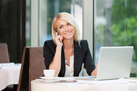 Beautiful blond businesswoman talking by phone. While sitting at cafÃ© with laptopの写真素材