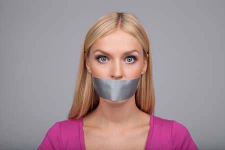 She will not gossiping anymore. Portrait of beautiful young women with her mouth covered with an adhesive tape standing isolated on grayの写真素材