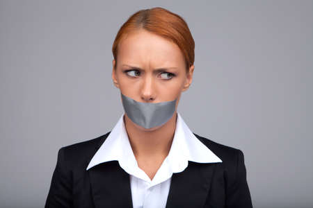 Gossip businesswoman. Portrait of beautiful young businesswoman with her mouth covered with an adhesive tape standing isolated on grayの写真素材