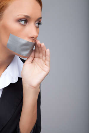 Businesswoman gossiping. Portrait of beautiful young businesswomen with her mouth covered with an adhesive tape standing isolated on gray and  holding her hand near mouthの写真素材