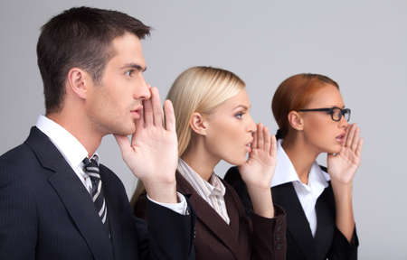 Global gossiping. Side view of three attractive young business people telling gossips standing with their hands covering mouth while isolated on grayの写真素材