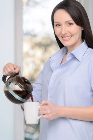 Want some coffee? Beautiful middle-aged woman standing in front of the window pouring some coffee to the cupの写真素材