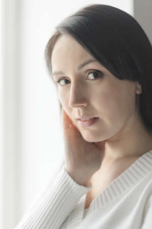 Woman standing in front of the window. Portrait of beautiful middle-aged woman standing in front of the window and holding her hand on the chiの写真素材