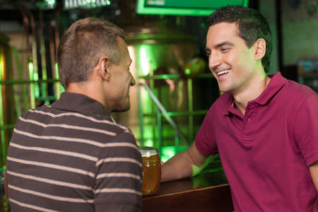 Friends having fun at bar. Two cheerful male friends talking at bar and drinking beerの写真素材