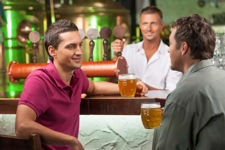 Talking at bar. Two cheerful male friends talking at bar and drinking beer while bartender serving beer at the backgroundの写真素材