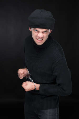 Busted burglar. Angry burglar in handcuffs grimacing at camera while standing against black backgroundの写真素材