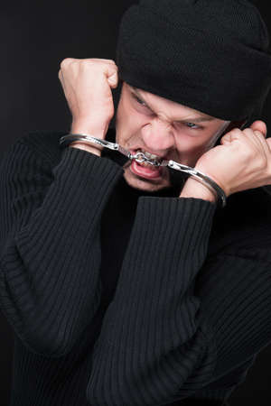 Trying to escape. Angry burglar in handcuffs grimacing at camera while standing against black backgroundの写真素材