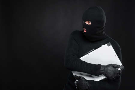 Stealing a suitcase. Frustrated thief in black balaclava holding a stolen suitcase while isolated on blackの写真素材