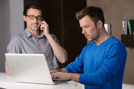 Working on project. Two cheerful businessmen standing near laptop while one of them talking on the phoneの写真素材