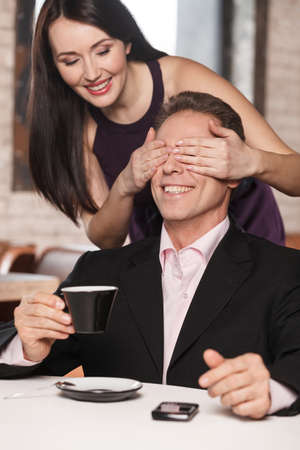 Guess who? Beautiful women closing her boyfriendï¿½ eyes while he is sitting with a cup of coffeeの写真素材