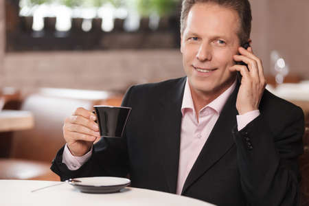 Talking to business partner. Cheerful businessman drinking coffee and talking at phone while sitting at restaurantの写真素材
