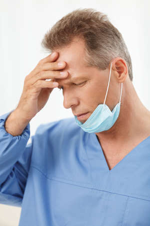 He made all possible to save the life. Depressed mature doctor in blue uniform standing with his hand in hairの写真素材