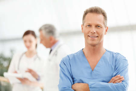 Only the best medical treatment. Portrait of a smart mature male doctor in blue uniform standing in front of his colleagues and smilingの写真素材