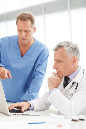 Discussing medical reports. Mature doctor using computer and discussing medical reports with his colleagueの写真素材