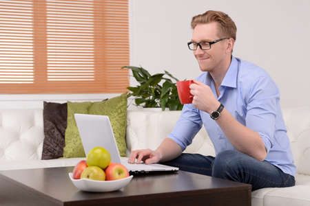 Relaxing at home. Cheerful young men in glasses drinking coffee and using computerの写真素材