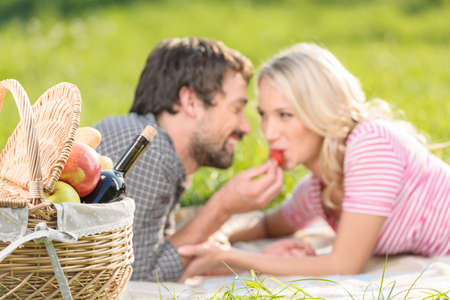 A strawberry for her. Young men feeding his girlfriend with strawberry lying on the grass in spring parkの写真素材