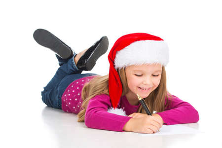 Beautiful little blond girl writing letter to Santa Claus. While laying isolated on white in Santa Claus hatの写真素材