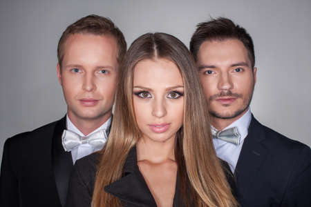 Portrait of three attractive people isolated over grey background. Sexy blond girl standing on foreground and stylish men in the same classical suites on backgroundの写真素材