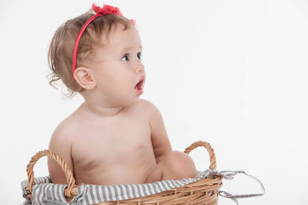 Cut little baby girl in red hoop sitting in basket. Looking away with surprise while isolated over white backgroundの写真素材