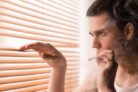 young handsome male looking through window. man smoking cigarette and watching from homeの写真素材