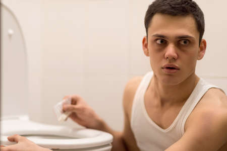 young handsome man sitting in toilet. drug addict alarmed and listening attentivelyの写真素材