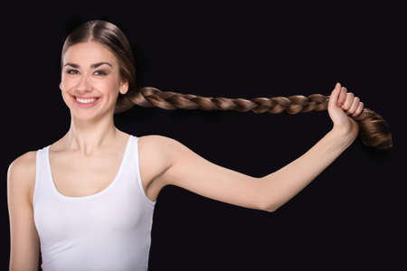 young woman pulling long plait. beautiful girl waist up holding hair on black backgroundの写真素材
