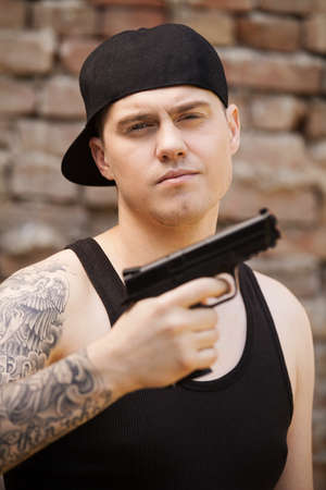young guy with tattoo holding gun. gangster looking into camera with brick wall backgroundの写真素材