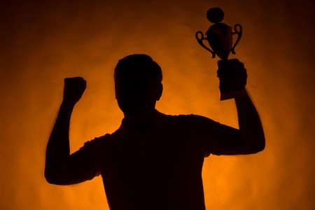 silhouette of man holding champion cup. waist up man standing on orange backgroundの写真素材