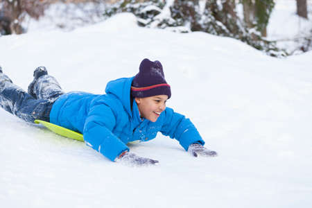 boy lying on sledges and sliding down hill. child speeding from hill on snow in parkの写真素材