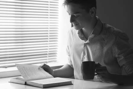 smiling guy sitting at table and reading book. black and white image of young man drinking teaの写真素材
