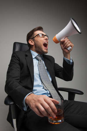 Young businessman shouting and sitting on chair. closeup of guy sitting with megaphone and glass on white backgroundの写真素材