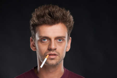 Young man smoking cigarette on black background. Handsome young man with brown hair wearing red t-shirtの写真素材