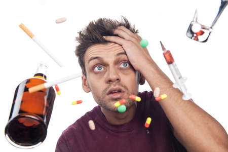 man sitting and holding head over table. sad guy with scattered pills, syringe, glass and bottleの写真素材
