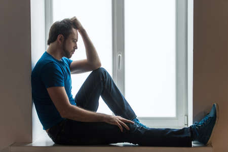 Handsome young man sitting on windowsill. Guy holding head and wearing blue shirt and jeansの写真素材