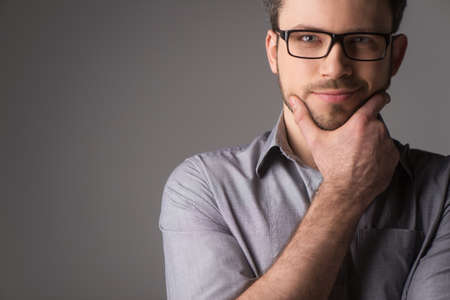 Close-up portrait of attractive young man holding chin. Man standing on gray background with glasses