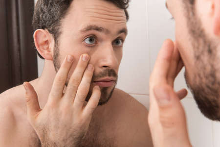 Closeup of young man examining his eye in mirror. Young man looks at his eye and thinkingの写真素材