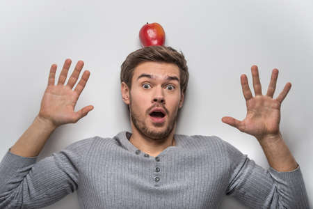 Handsome young guy balancing red apple on his head. guy wearing grey sweater isolated on white background の写真素材