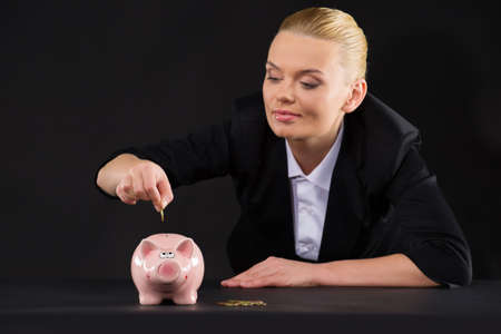 Pink piggy bank standing on dark table. closeup of woman putting coin inside piggy bankの写真素材