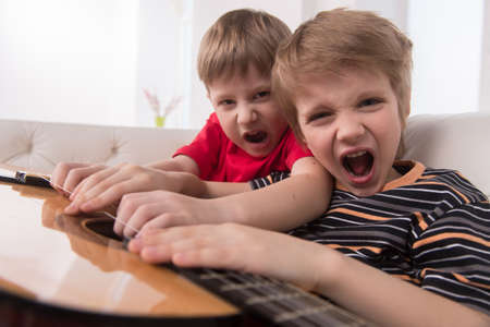 two caucasian boys playing acoustic guitar. boy sitting on couch and playing loud musicの写真素材